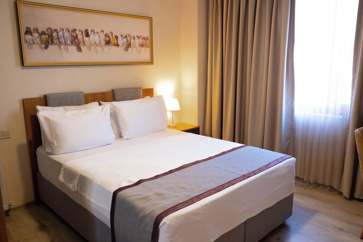 A neatly made bed with white linens and a decorative border, a bedside lamp, a framed artwork above the bed, and a window with curtains.