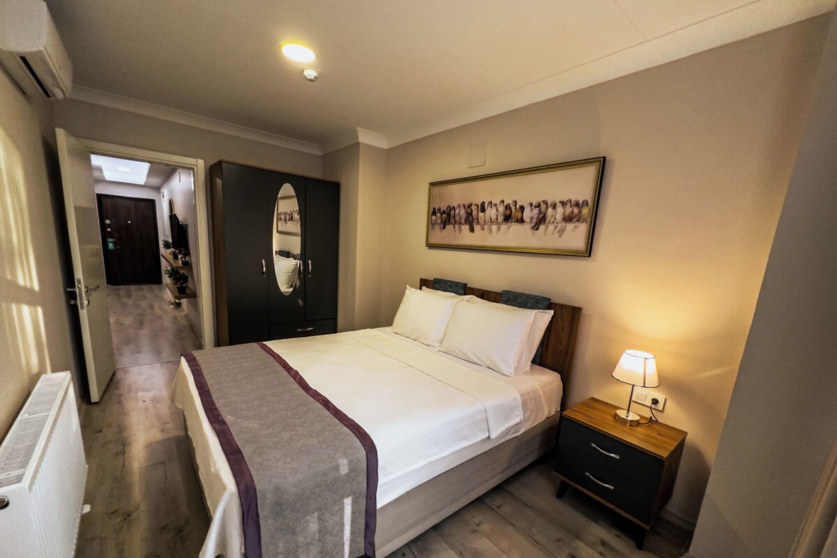 Habitación de hotel con cama, mesita de noche, lámpara, armario y un pasillo que conduce a otra habitación.