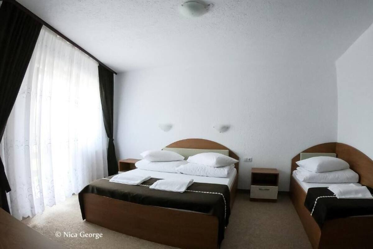 A hotel room with two beds, a large window with curtains, a bedside table, and a wall-mounted light fixture.