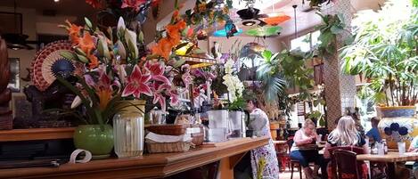 A restaurant with a variety of plants and flowers decorating the interior, wooden furniture, and a tiled floor.