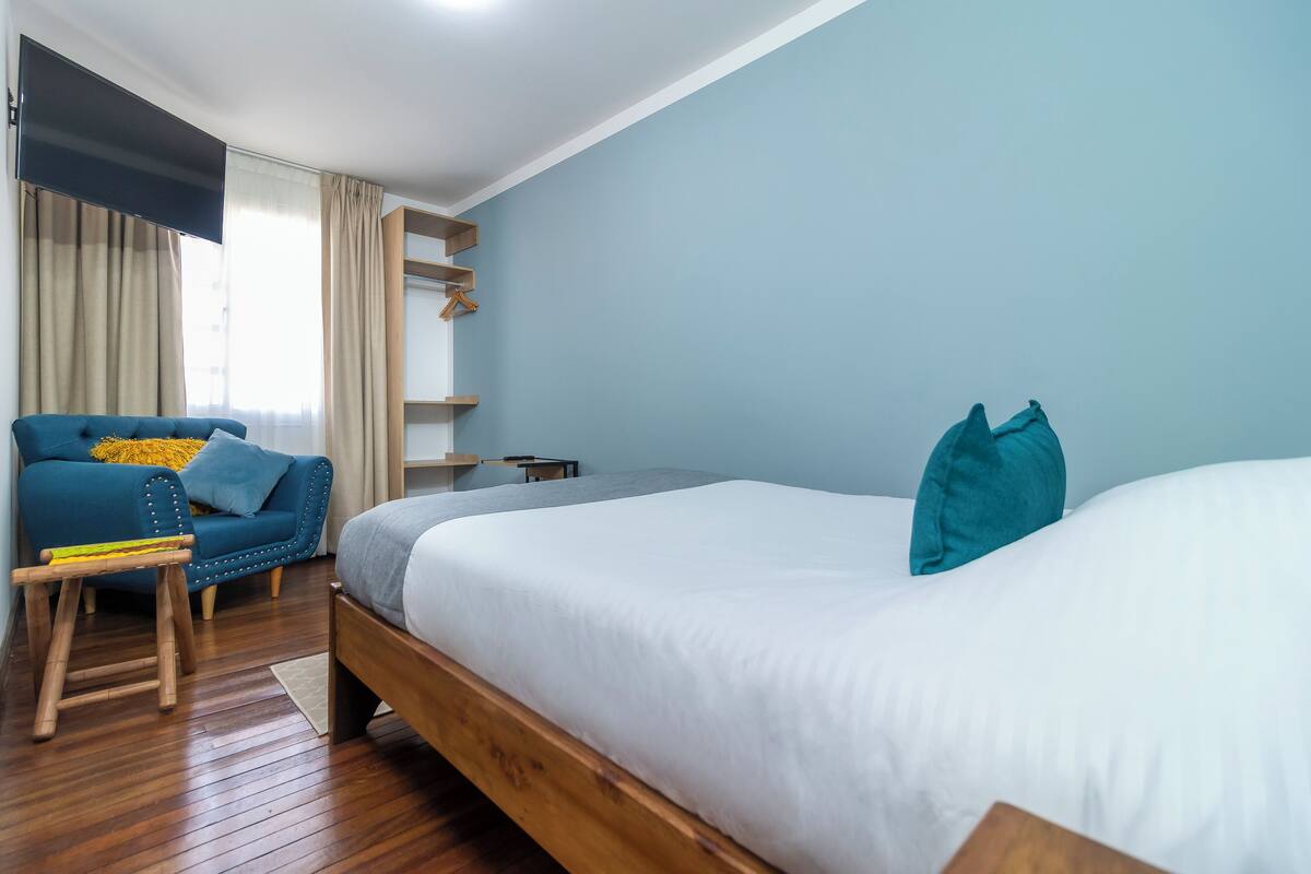 A bedroom with a large bed, a blue armchair, a wooden stool, and a TV mounted on the wall.