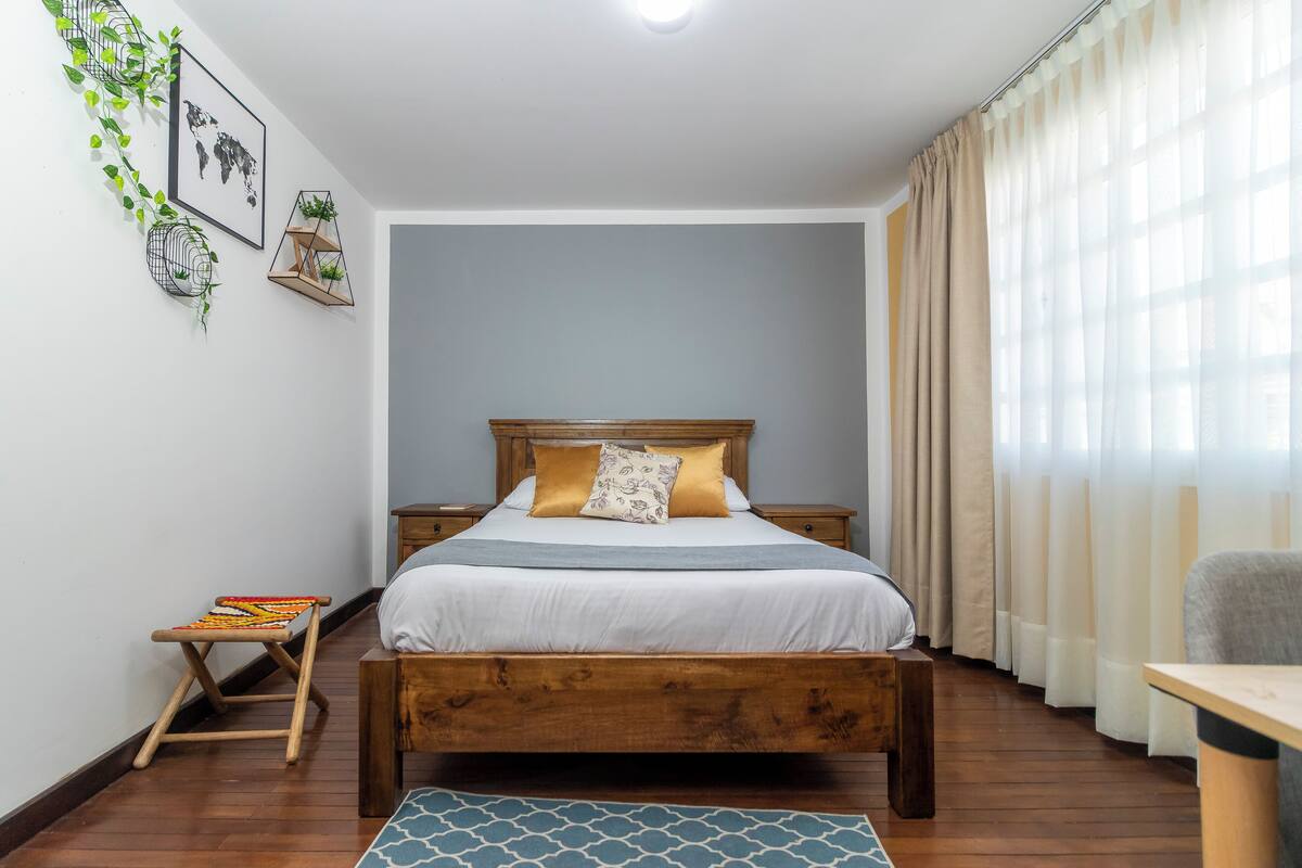 A bedroom with a wooden bed, a small bedside table, a chair, a patterned rug, and a world map on the wall.