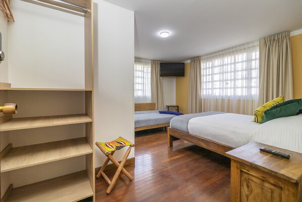 A hotel room with a wooden floor, a bed with a white mattress, a small table with a remote control, a wooden shelf unit, and a window with curtains.
