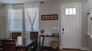 A dining area with a wooden table and chairs, a window with white curtains, a door, and a wall-mounted shelf with a plant and a decorative item. - Family home at Rohnert Park (Rohnert Park)