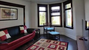 Un salon avec un canapé rouge, un tapis à motifs, une table à manger avec des chaises et de grandes fenêtres équipées de stores.