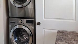 Une buanderie moderne et compacte, dotée de deux lave-linge et deux sèche-linge à chargement frontal en acier inoxydable, d’une porte blanche et d’un plan de travail en granit.