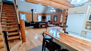 Een interieur van een blokhut met een houten trap, een zithoek met een bank en salontafel, een keuken met een koelkast, en een eetgedeelte met stoelen en een tafel.