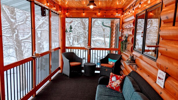 Een gezellig houten blokhutinterieur met een houten terras, rieten meubels en grote ramen met uitzicht op sneeuw.