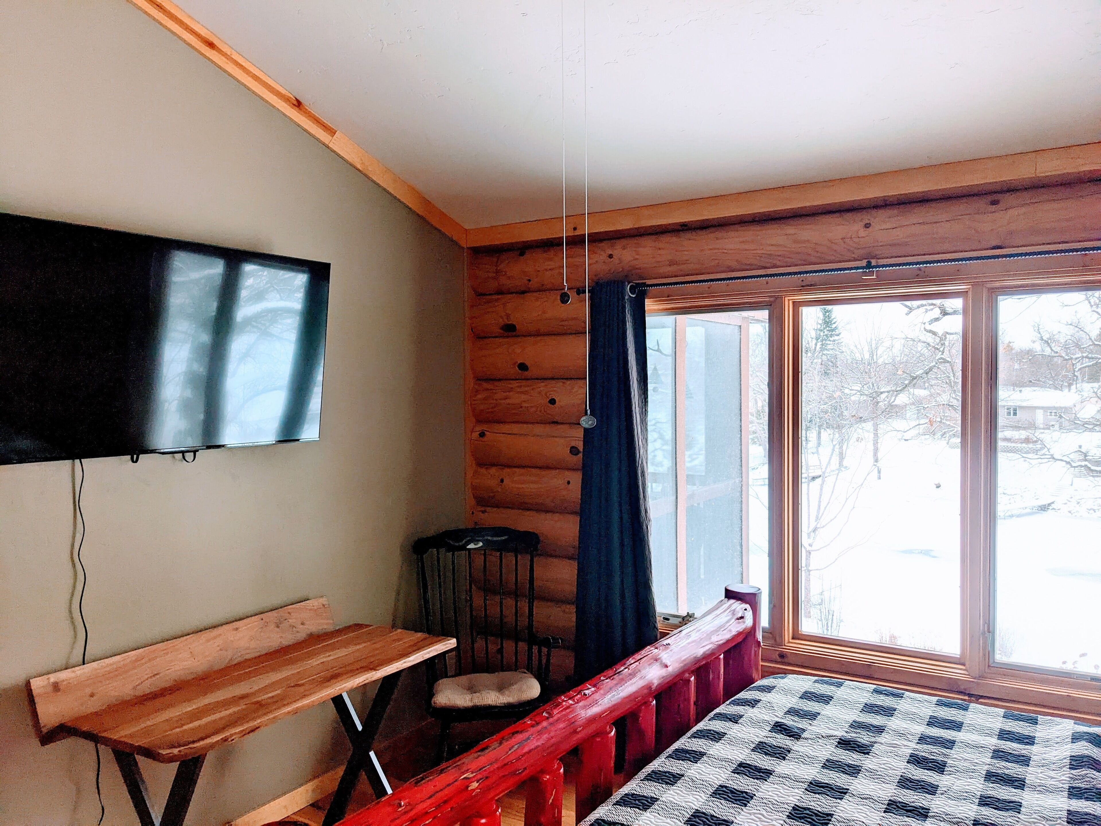 A cozy cabin room with a wooden bench, a bed with a checkered quilt, a television mounted on the wall, and a window with curtains.
