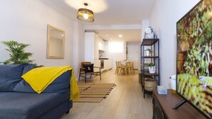 Ein modernes Wohnzimmer mit Sofa, Couchtisch, Essbereich mit Stühlen, einer Küche im Hintergrund und einem Fernseher auf einem Schrank.
