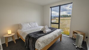 A bedroom with a bed, a bedside table with a lamp, a radiator, and a large window with a view of the outdoors. - Southern Stars - FREE WiFi (Twizel)