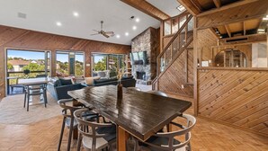 A spacious living area with a large wooden dining table, comfortable seating, a fireplace, and large windows offering a view of the outdoors. - Lake Travis Gem, Golf, boat, Tennis, Pool (Point Venture)