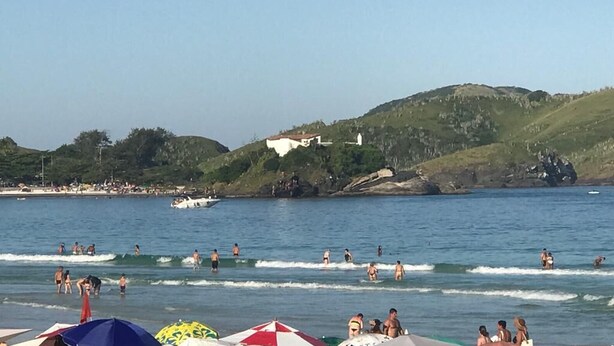 A beach with people swimming and sunbathing, a boat in the water, and a hill with buildings in the background. - Suíte Palmeiras  (Cabo Frio)