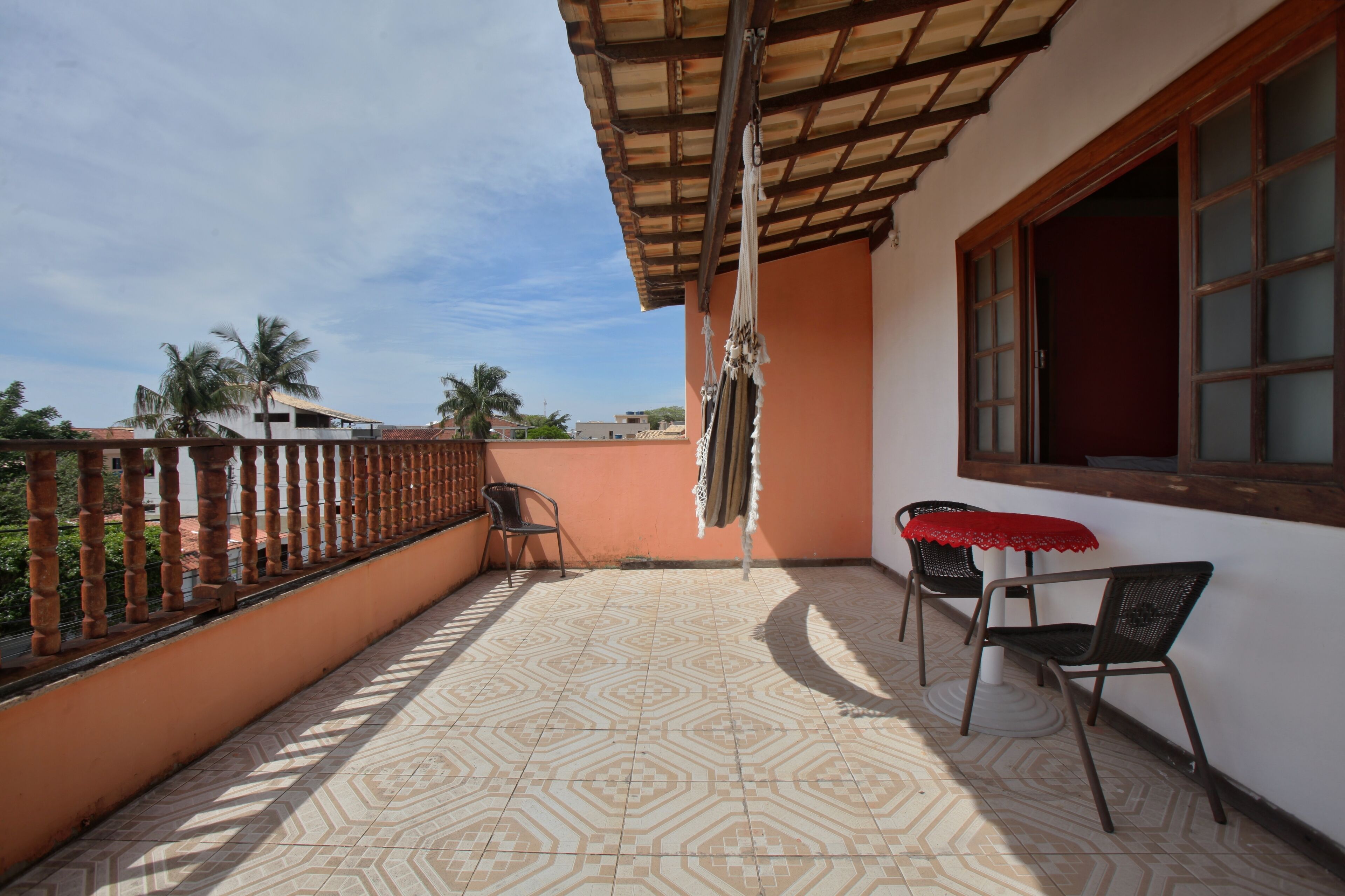 Un balcón con baldosas estampadas, baranda de madera, dos sillas y una mesa con mantel rojo.