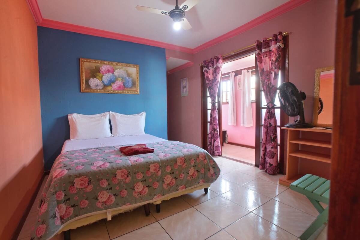 A bedroom with a bed, a ceiling fan, a window with curtains, and a small table.