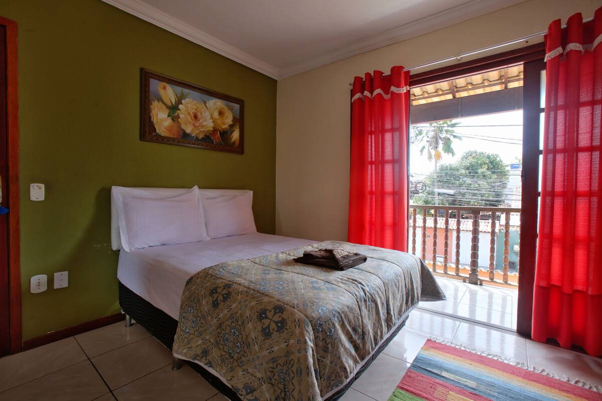 A bedroom with a bed, a painting on the wall, and a balcony with red curtains.