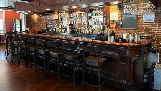 A bar area with a wooden bar counter, multiple stools, and a variety of bottles and glasses. - Publick House Hotel Inc. (Chester)
