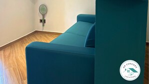 A teal sofa, a flat-screen TV, an air conditioner, a wall-mounted decorative piece, and a logo on a teal pillar. - Renovated 37m2 studio located 30 meters from Anse à L'âne beach (TROIS ILETS)