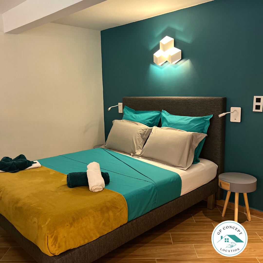 Ein modernes Schlafzimmer mit einem Bett, türkis-beigen Bettwäsche, einem kleinen Nachttisch und einer Wandleuchte.
