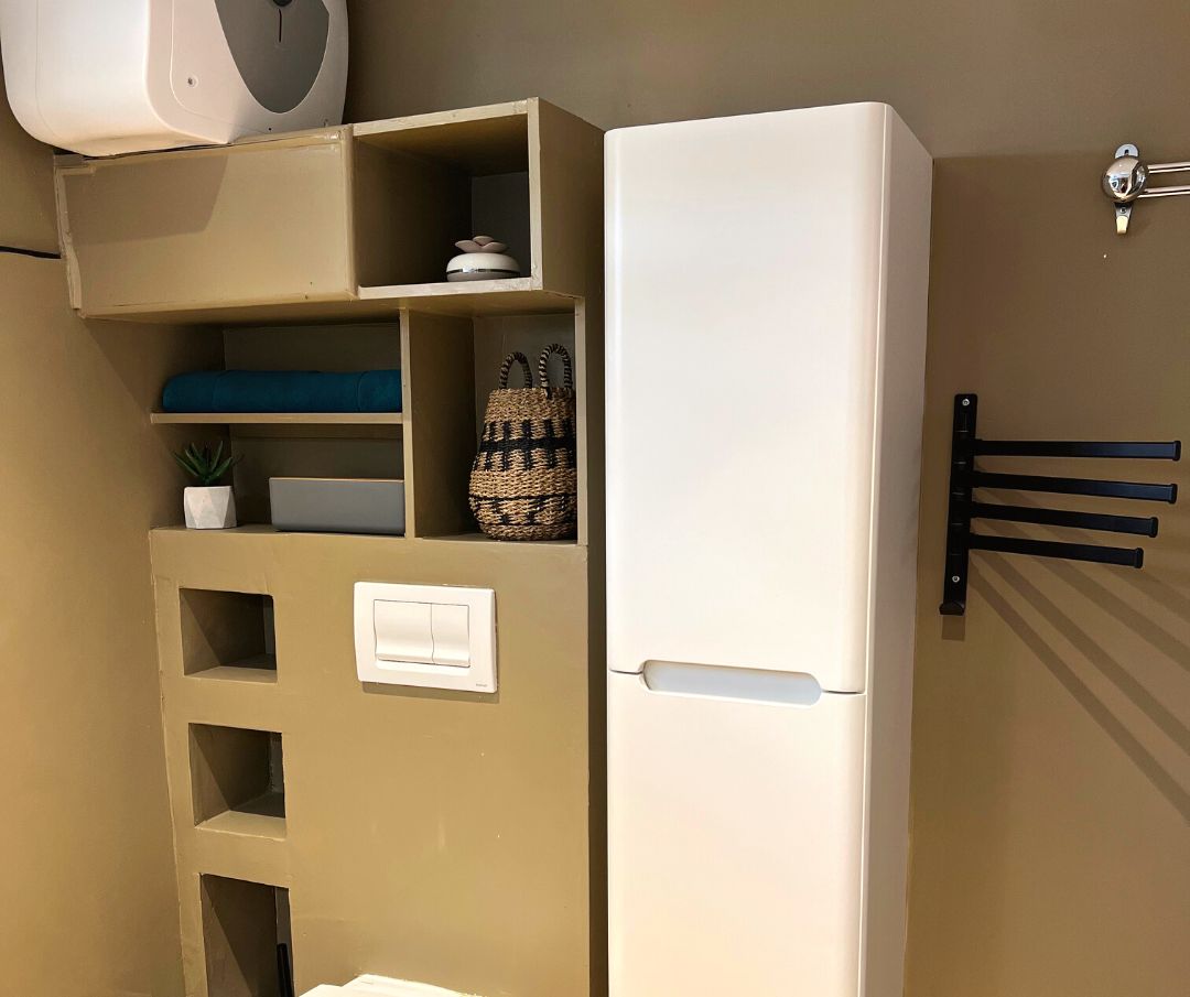 A compact bathroom with a white toilet, a built-in sink, a shelf with a potted plant, and a towel rack.