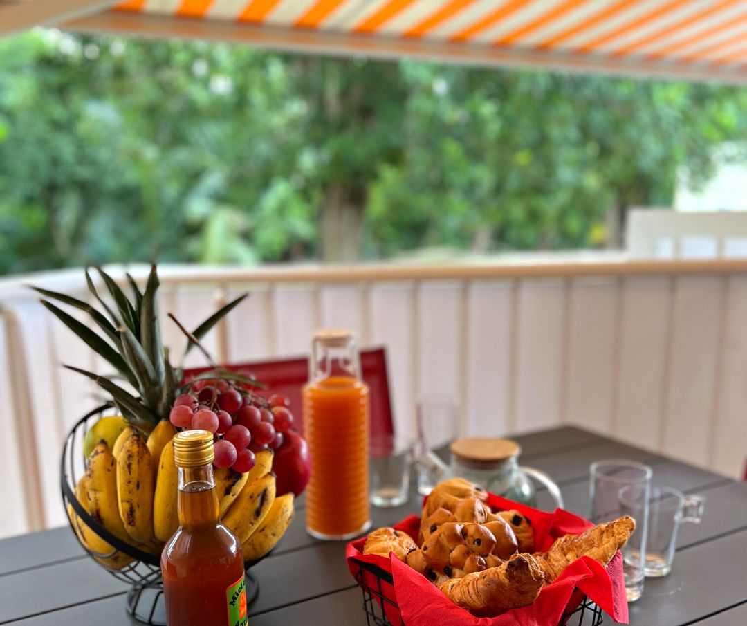 A table with a fruit bowl, a bottle of sauce, and a basket of pastries on an outdoor patio.