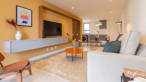A modern living room with a flat-screen TV, a sofa, a coffee table, and a dining area in the background. - Tabas | Moderno apto em Ipanema - RJ | IP0019 (Rio de Janeiro)