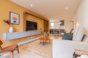 A modern living room with a flat-screen TV, a sofa, a coffee table, and a dining area in the background. - Tabas | Moderno apto em Ipanema - RJ | IP0019 (Rio de Janeiro)