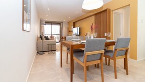 A modern dining area with a wooden table and grey upholstered chairs, a television, and a sofa in the background. - Tabas | Moderno apto em Ipanema - RJ | IP0019 (Rio de Janeiro)