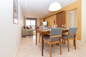 A modern dining area with a wooden table and grey upholstered chairs, a television, and a sofa in the background. - Tabas | Moderno apto em Ipanema - RJ | IP0019 (Rio de Janeiro)