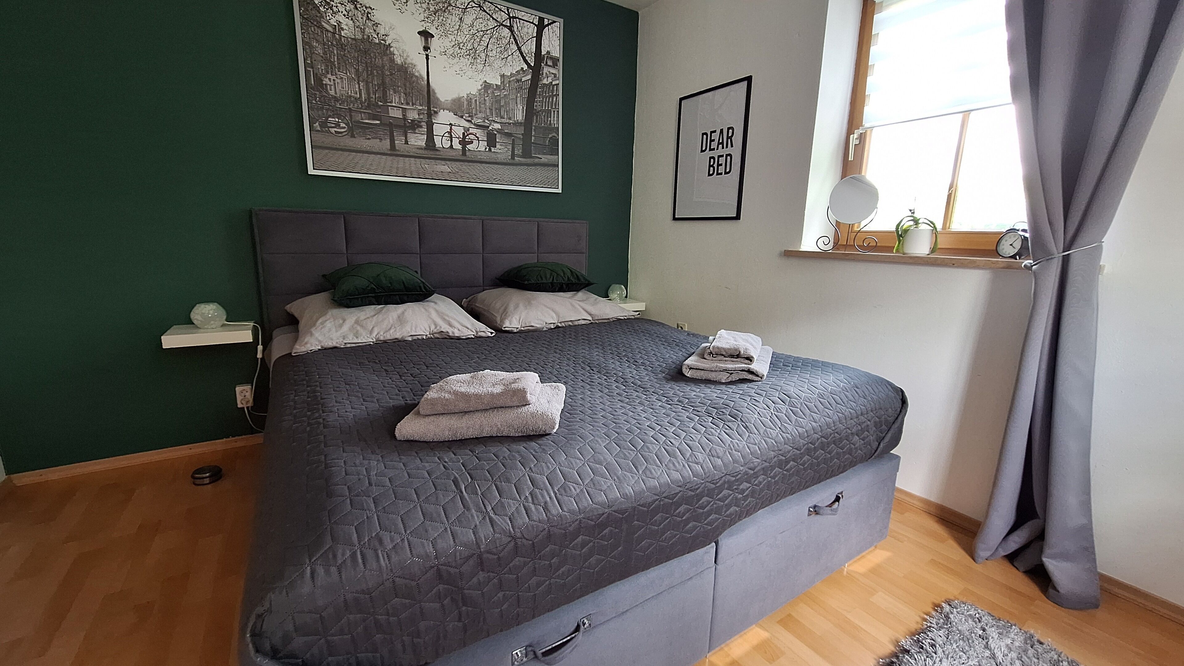 Une chambre à coucher avec un lit, des oreillers, une table de chevet, une fenêtre avec des rideaux, une horloge murale et un tableau encadré.