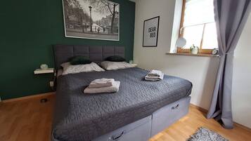 Une chambre à coucher avec un lit, des oreillers, une table de chevet, une fenêtre avec des rideaux, une horloge murale et un tableau encadré.