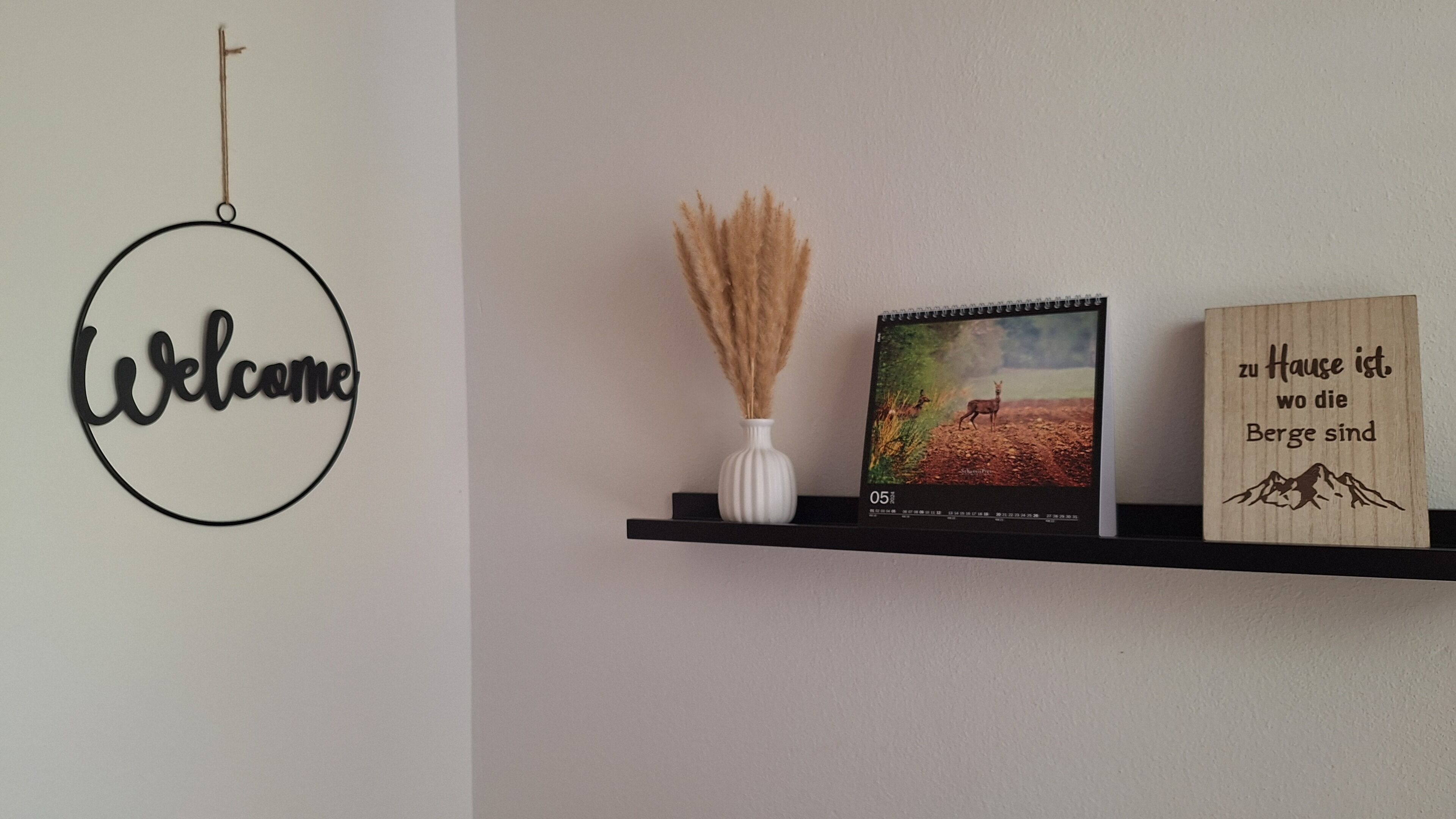 A wall with a 'Welcome' sign, a white vase with dried plants, a calendar, and a wooden sign with a mountain image.