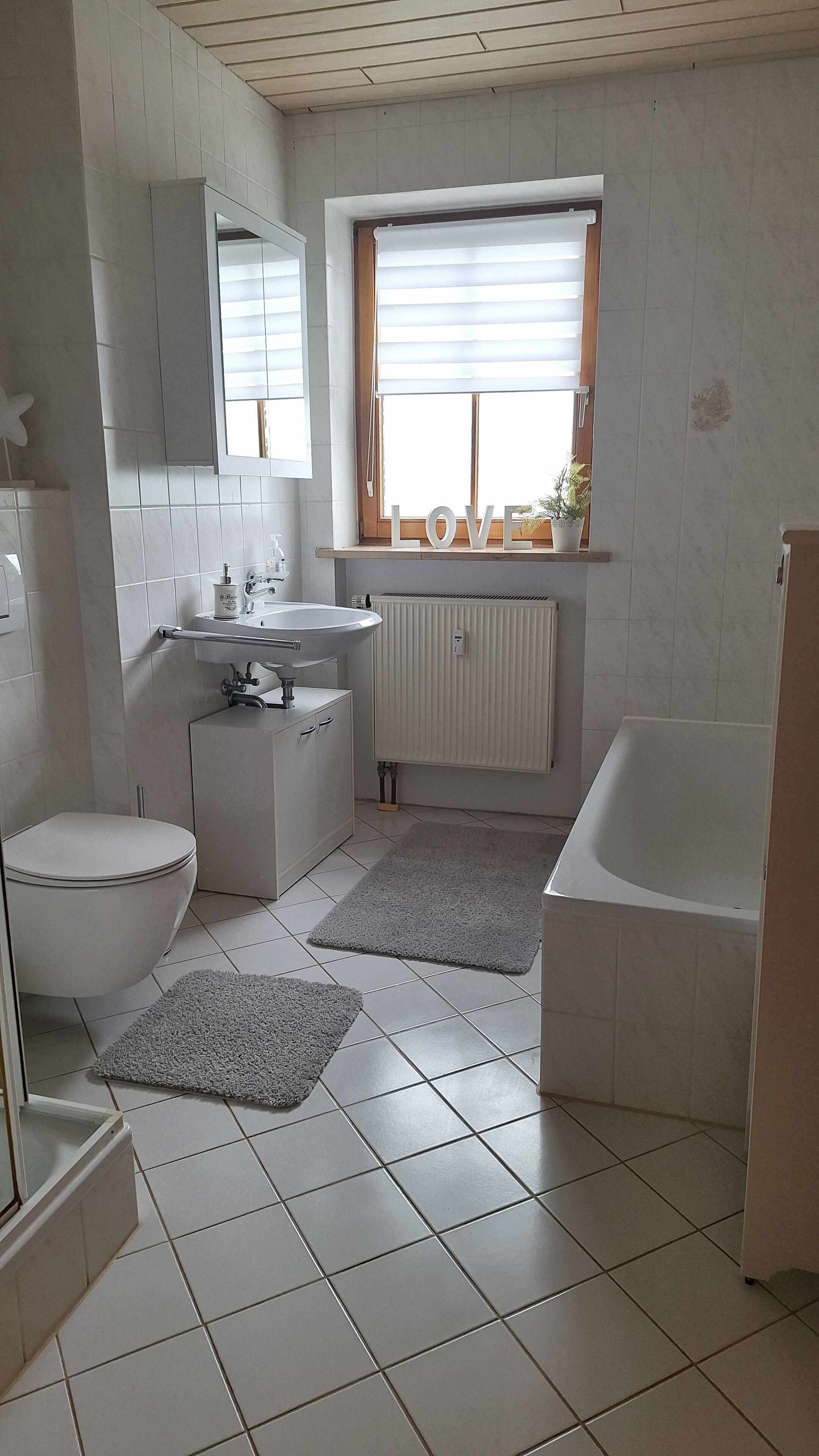 Ein modernes Badezimmer mit Toilette, Waschbecken und Badewanne. Ein Fenster mit Jalousien und ein Heizkörper sind vorhanden.