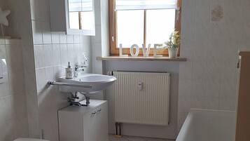 Ein modernes Badezimmer mit Toilette, Waschbecken und Badewanne. Ein Fenster mit Jalousien und ein Heizkörper sind vorhanden.