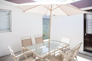 A glass-top table with four chairs, an umbrella, and a window with a view of a green fence.