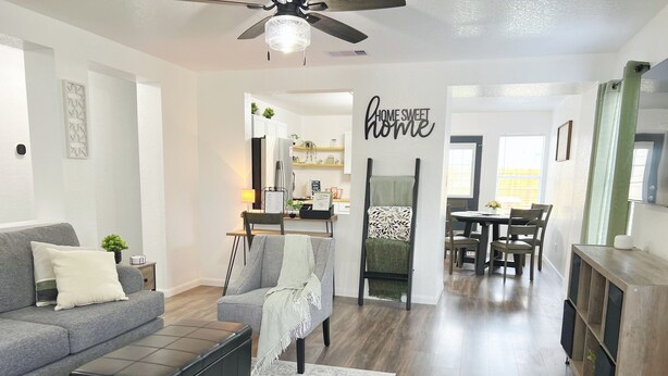 A modern living room with a grey sofa, black ottoman, and a patterned rug. A dining area with a table and chairs is visible in the background. - Relaxing 3BR w/ Waffle Bar•Lackland BMT•Seaworld (San Antonio)