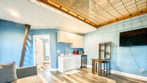 A modern kitchen with white cabinets, a dining area with a wooden table, and a living room with a gray sofa and a cowhide rug.