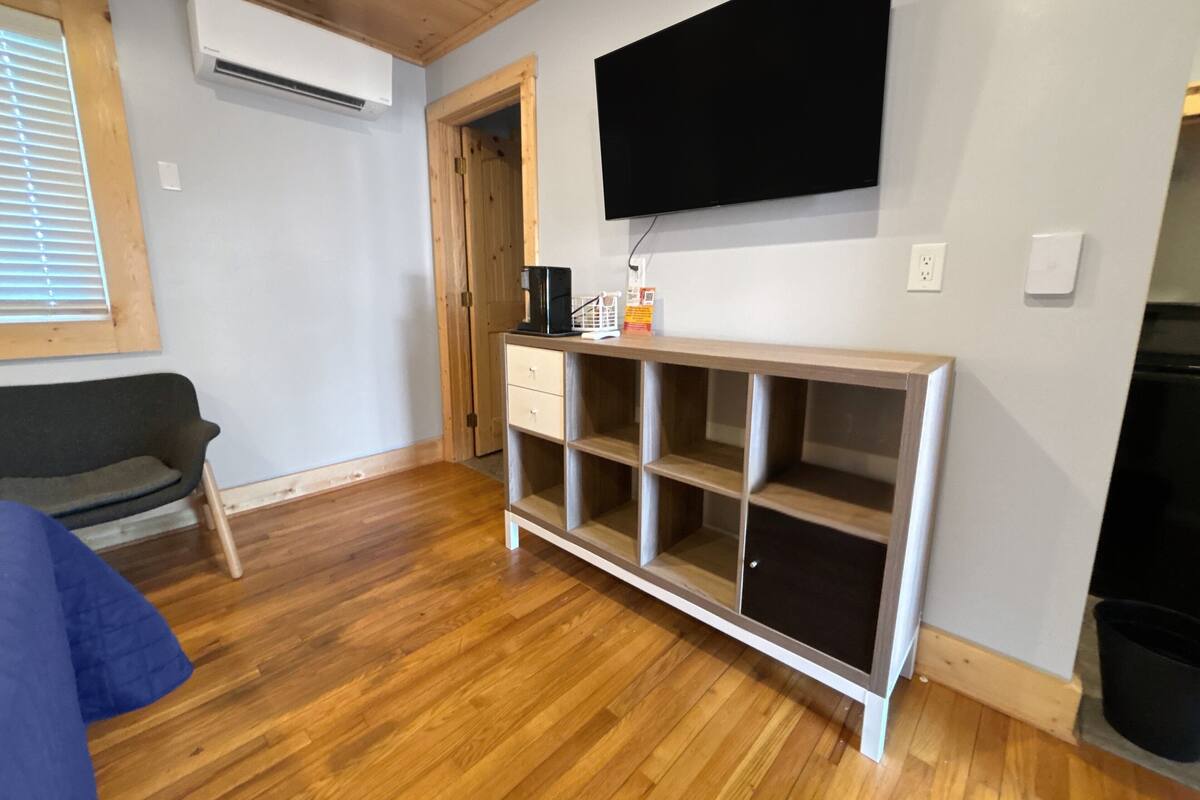 A room with a wooden floor, a sofa, a TV mounted on the wall, a coffee machine, and a small cabinet.