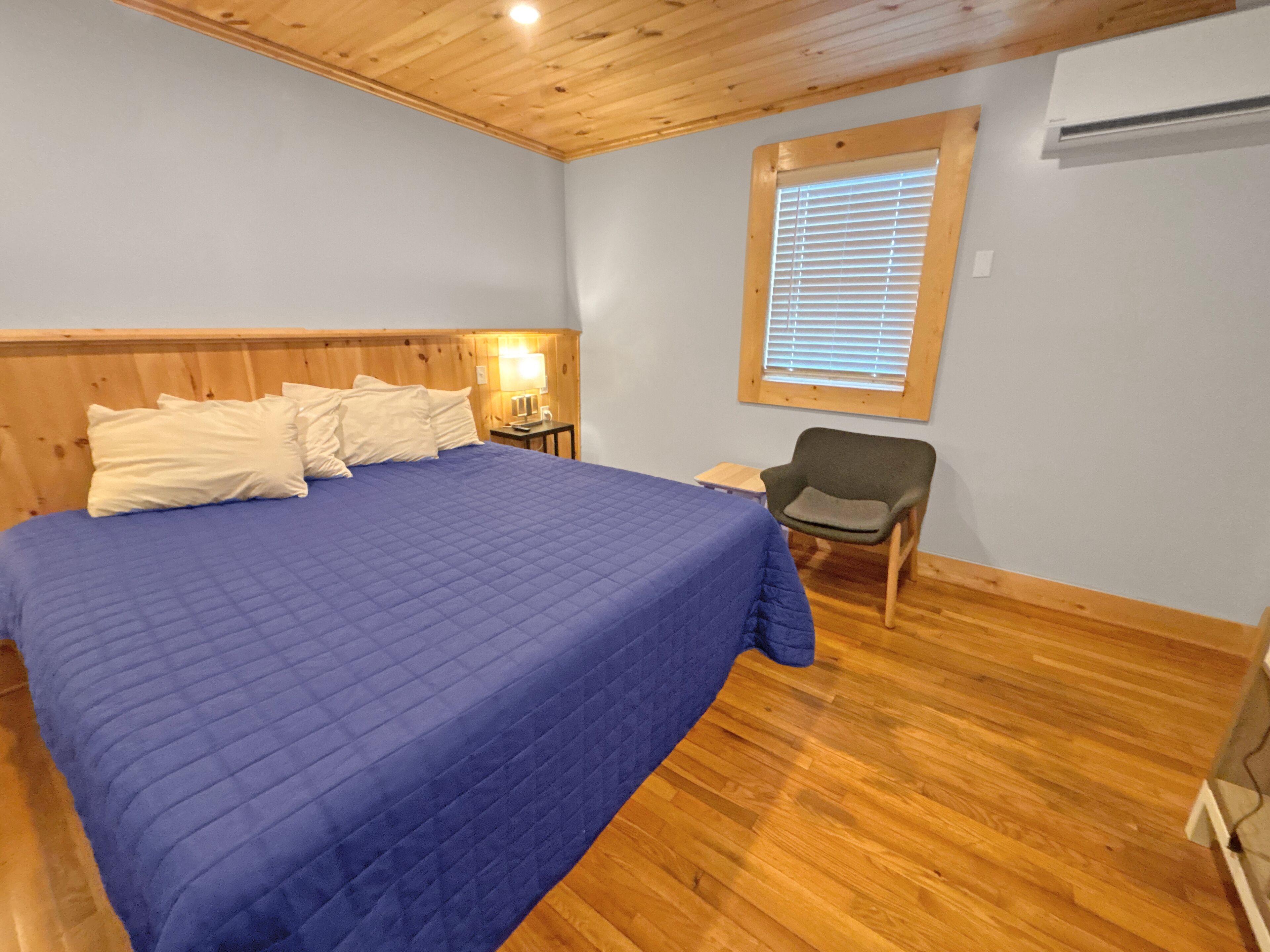 Une chambre à coucher avec une tête de lit en bois, une couvre-lit bleue, une chaise, une table de chevet et une fenêtre avec des stores.