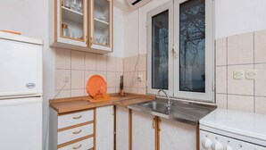 Een kleine keuken met witte kastjes, een inbouwoven, een spoelbak en een raam met uitzicht op kale bomen.