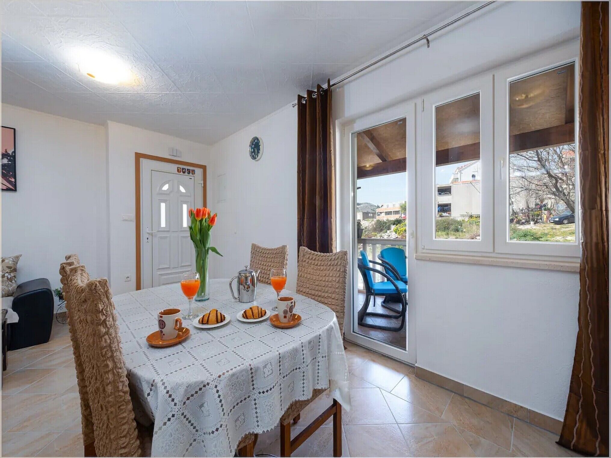 A dining area with a table set for two, a vase with flowers, and a view of the outside through a window.