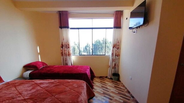 Ein Zimmer mit zwei Betten, einem fensterbelaubten Fenster, einem an der Wand montierten Fernseher und einem gemusterten Boden.