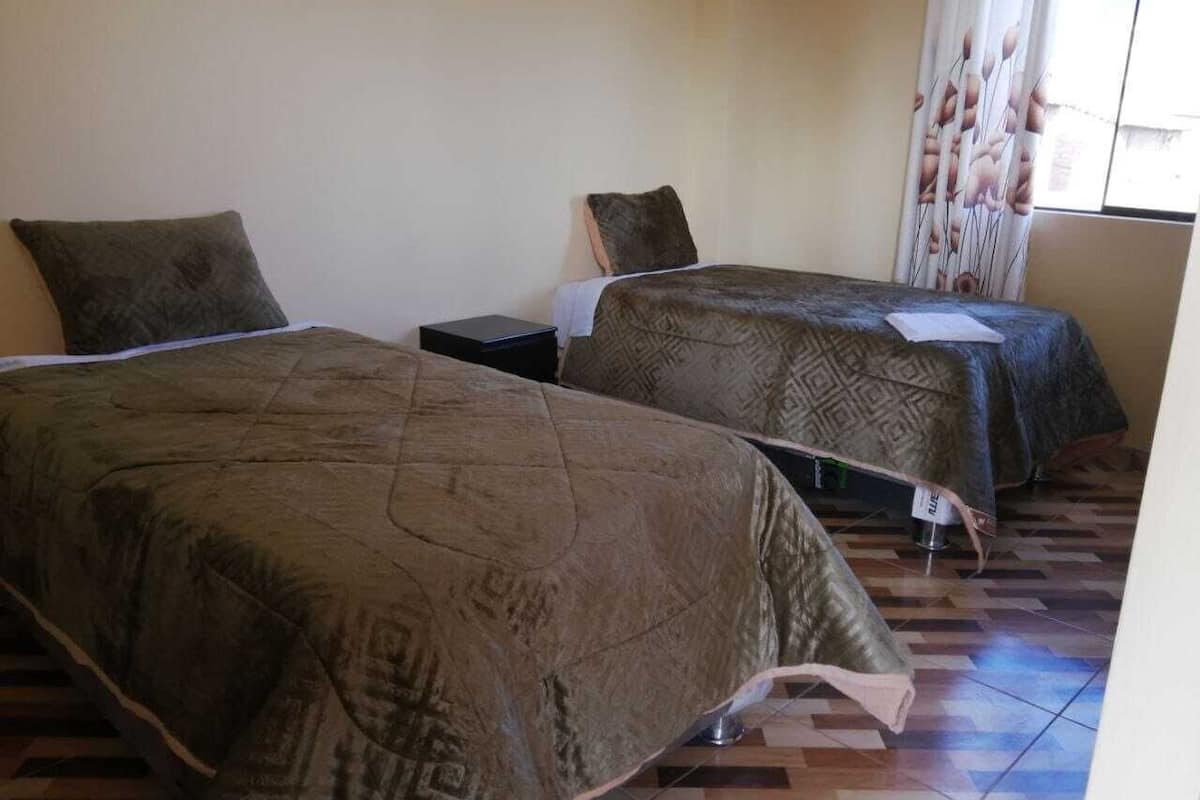 Habitación de hotel con dos camas, cada una con colcha y almohadas estampadas. El piso es de baldosas y hay una ventana con cortinas.