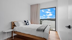 Een moderne slaapkamer met een houten bed, een witte nachtkastje en een groot raam met uitzicht op een meer en heuvels.