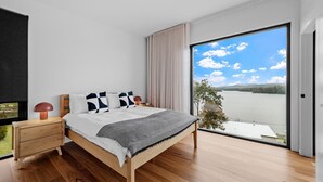 A modern bedroom with a wooden bed, a nightstand, a lamp, and a view of a lake through a large sliding glass door. - Piccolo Segreto Lake Conjola (Lake Conjola)