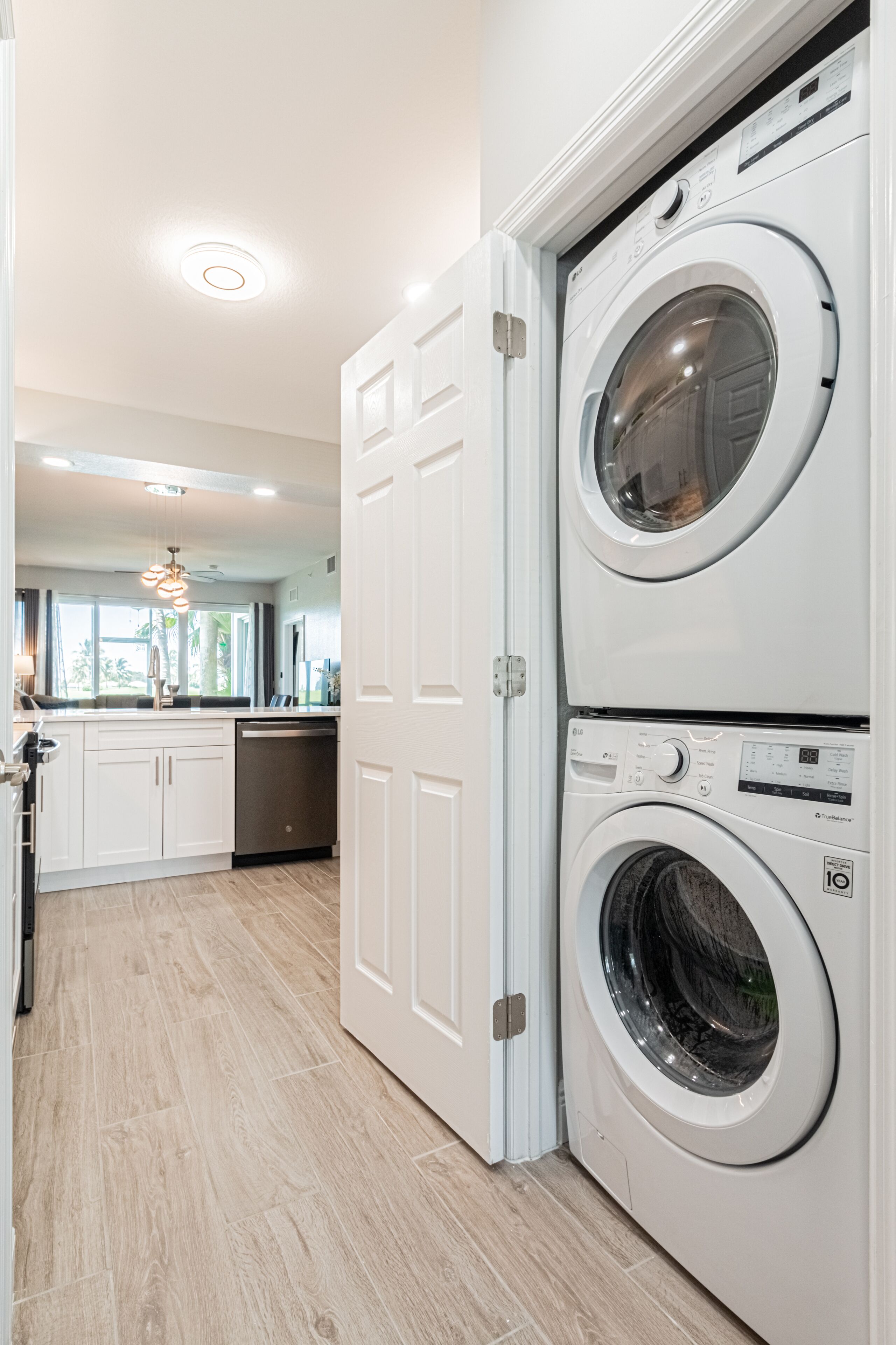 Une buanderie avec des lave-linge et des sèche-linge superposés, un coin cuisine équipé d’un lave-vaisselle, et une porte donnant sur une autre pièce.