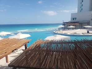 A poolside area with a view of the ocean, lounge chairs, umbrellas, and a building with a white facade. - Amazing Studio by the beach (Cancún)