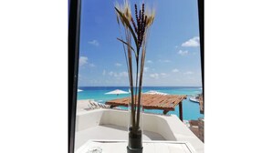 Blick von einem Balkon mit blauem Himmel und Meer. Ein Holztisch und Stühle stehen bereit, dazu eine Glasvase mit Trockenpflanzen.