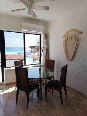 A dining area with a glass table and chairs, a ceiling fan, and a view of a beach and pool. - Amazing Studio by the beach (Cancún)