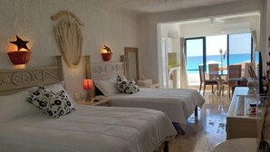 Ein Hotelzimmer mit zwei Betten, einem Deckenventilator, Blick auf den Strand und einer Essecke mit Tisch und Stühlen.